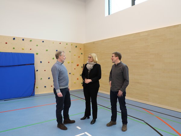 Schulleiter Carsten Böing, Oberbürgermeisterin Katharina Pötter und Holger Diekmann, Bauleiter vom städtischen Eigenbetrieb Immobilien- und Gebäudemanagement stehen in der Sporthalle vor der Boulderwand und unterhalten sich